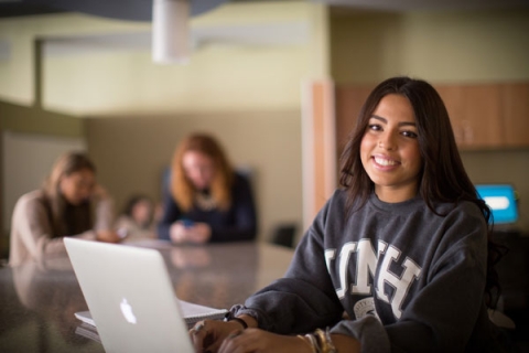 A student in a UNH sweatshirt smiles in front of her laptop.
