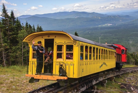 A train engineer waves from the back of a yellow passenger car going up a mountain